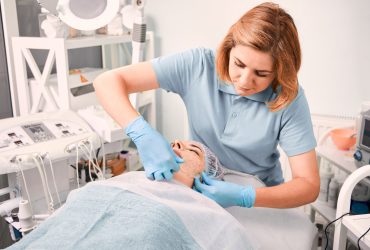 Cosmetologist in sterile gloves removing deep cleansing mask from female client face. Woman having skincare procedure at wellness medical center. Concept of beauty and facial treatment.